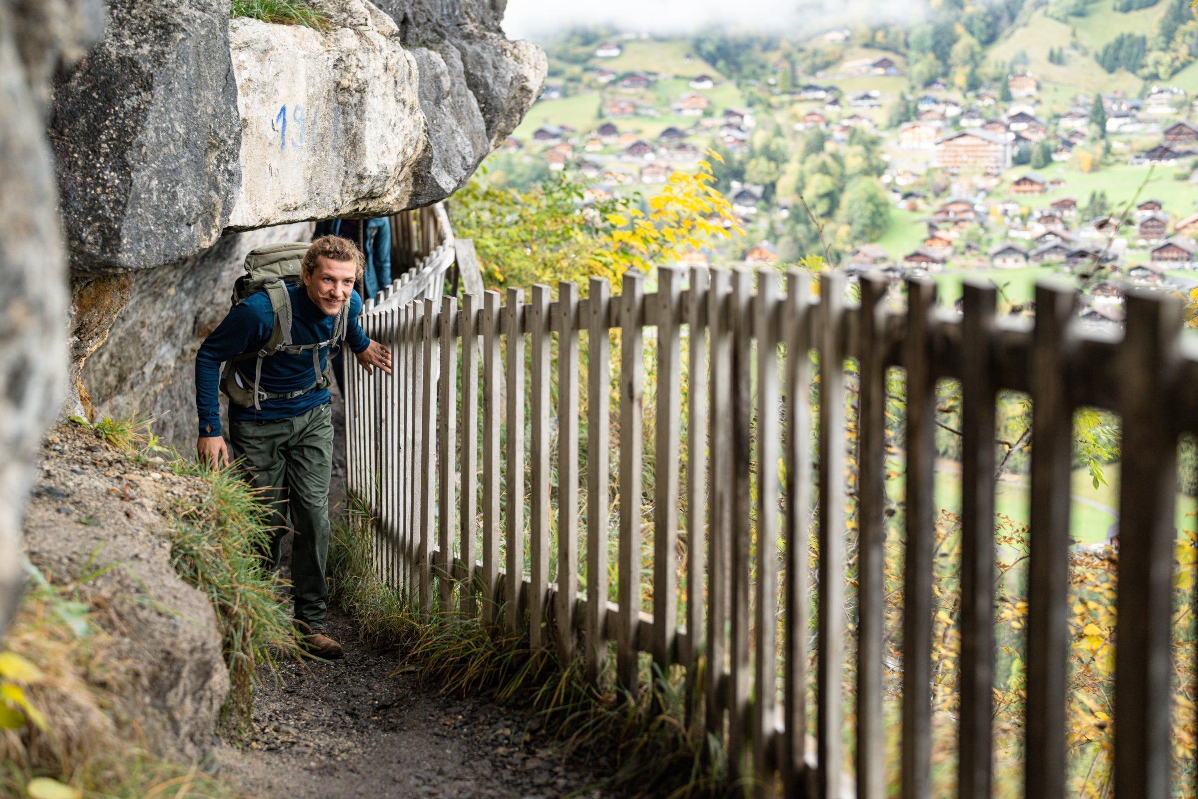 Hiker navigating a narrow trail with a wooden fence and scenic village view