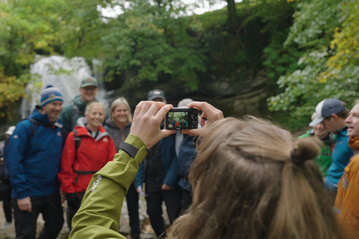 A group of people outdoors in front of a waterfall and trees, with a woman in the foreground holding a digital camera