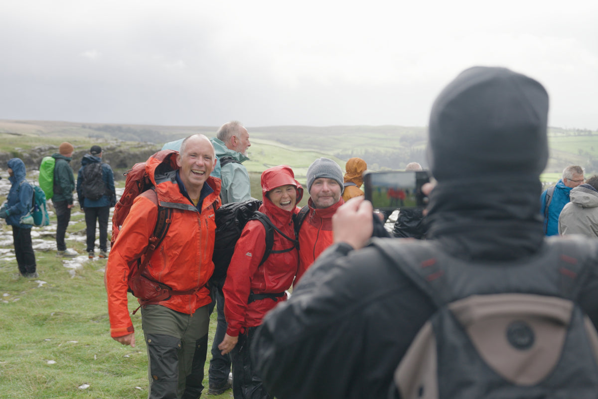 A group of hikers posing outdoors on a grassy hill under an overcast sky, with one person in the foreground taking a photo