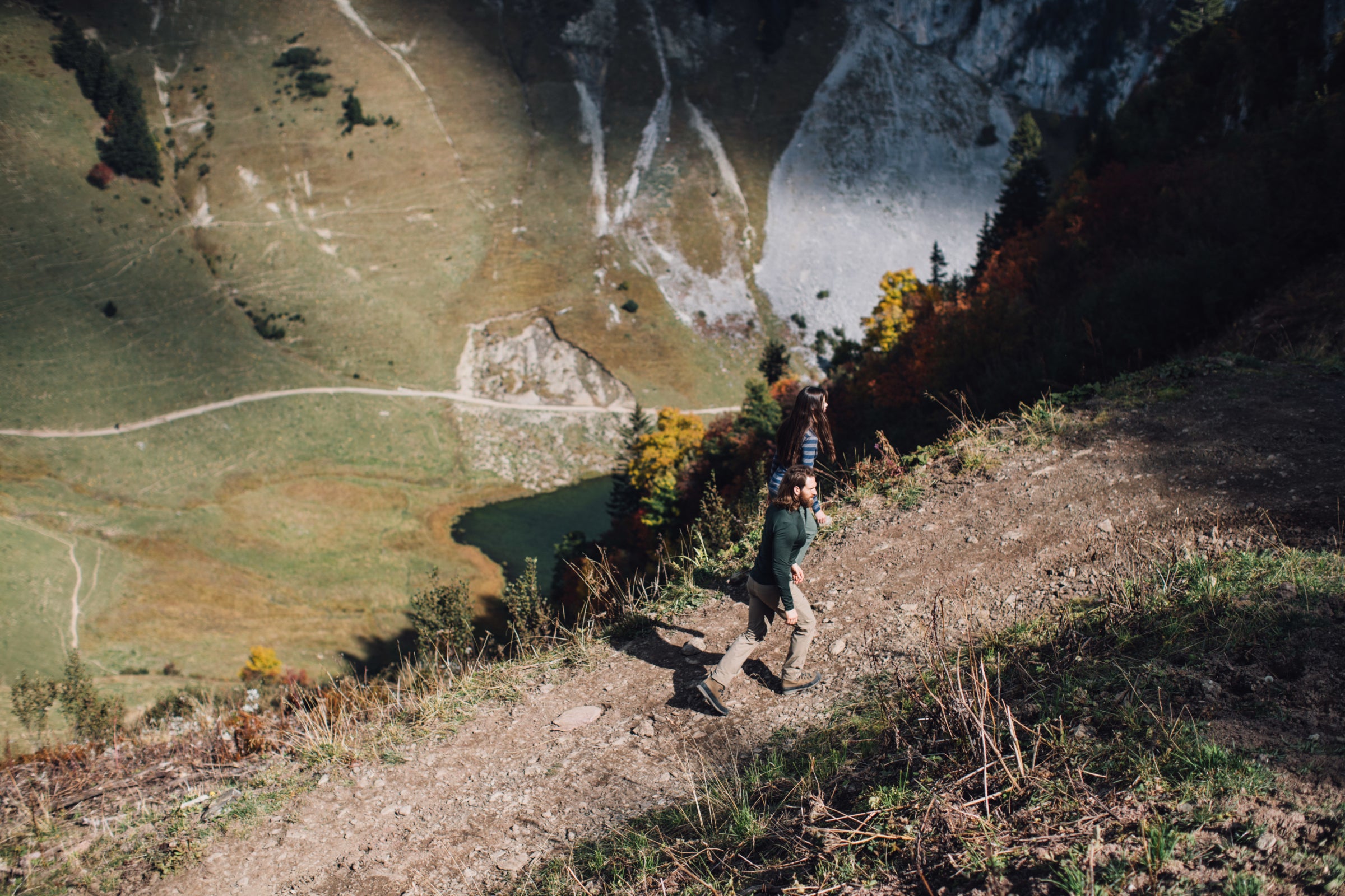 Deux randonneurs qui descendent un sentier de montagne raide avec vue sur un lac et des chemins en contrebas dans la vallée.