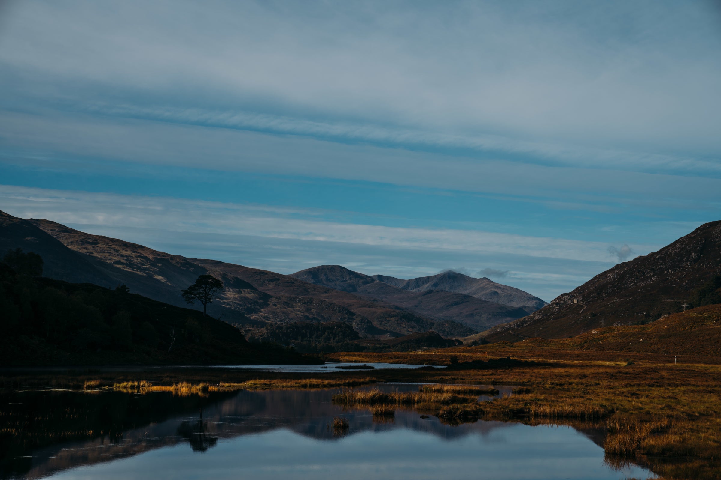 Un lac calme reflétant montagnes et ciel bleu aux nuages délicats, entouré de bruyère et d’herbes.