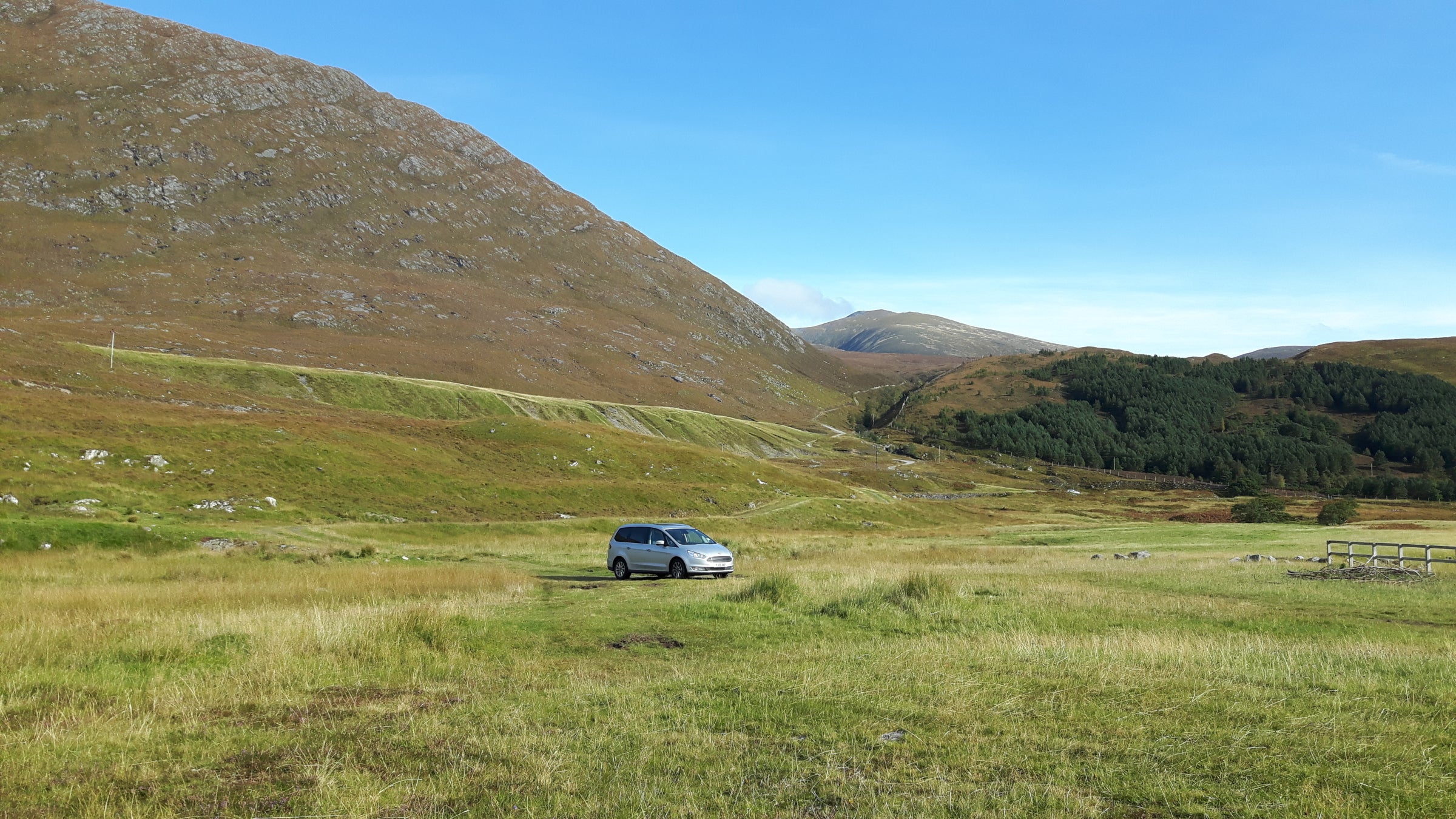 A silver car parked in a vast grassy field with large hills in the background