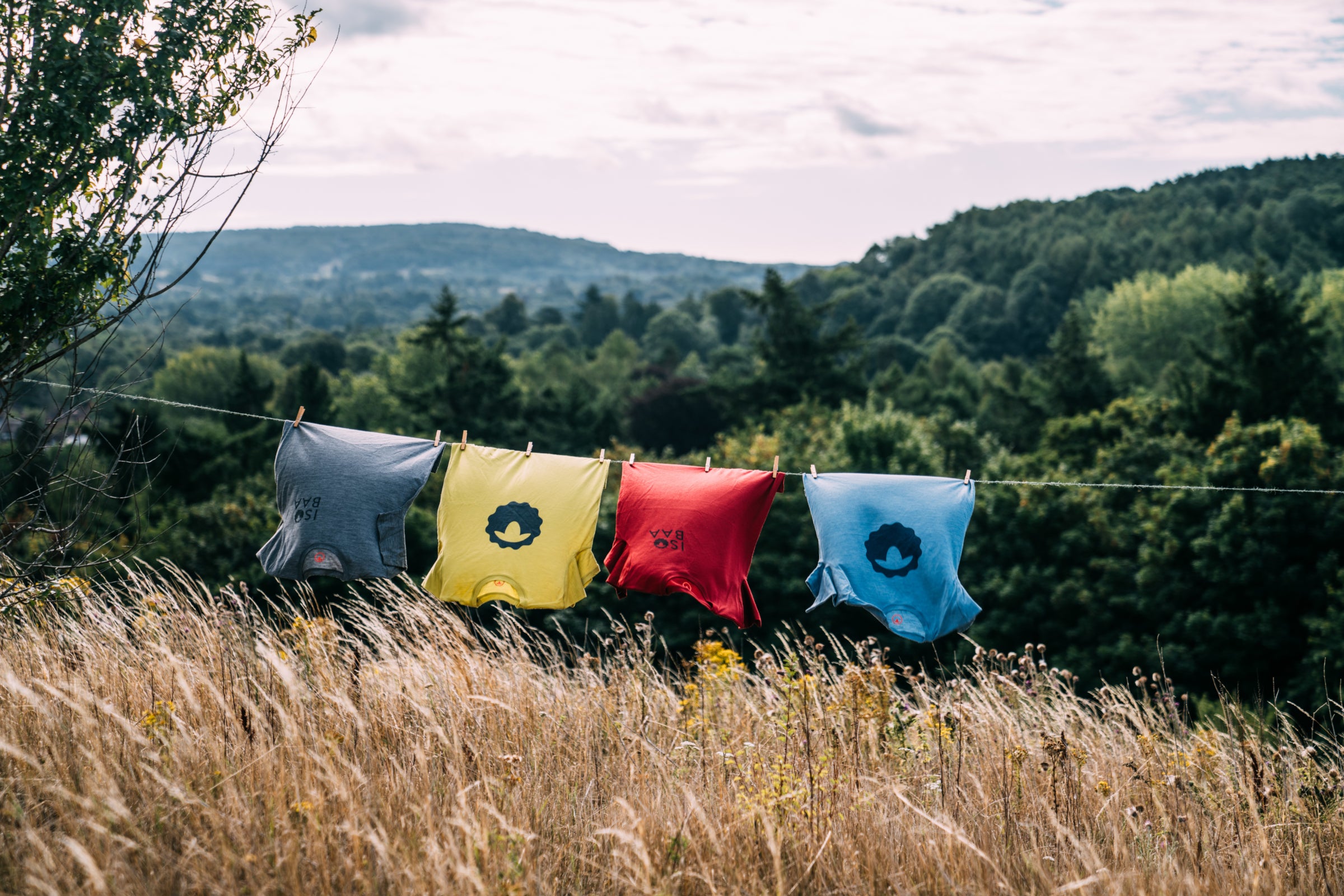 Vier farbenfrohe Merino Wool T-Shirts mit Isobaa-Logoprint hängen auf einer Wäscheleine in einem sonnigen Feld, mit Bäumen und Hügeln im Hintergrund.